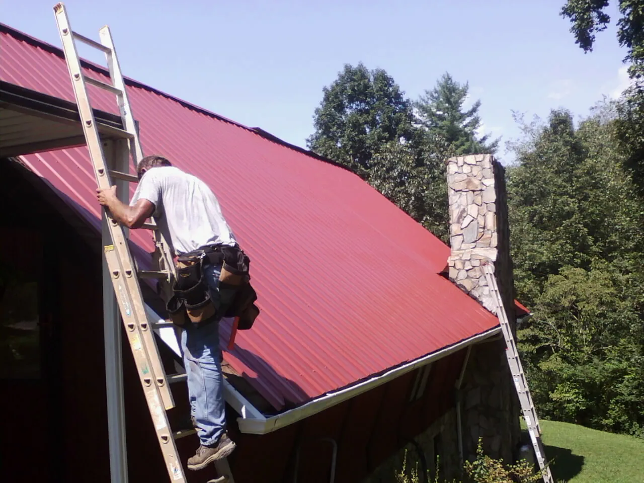 Roofer on a ladder beside a red metal roof and stone chimney — Soffit & Fascia Repair by Roofing Brothers in Steamboat Springs